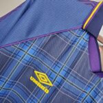 Close-up of blue plaid fabric with purple edging and a yellow embroidered logo/word along the bottom edge.