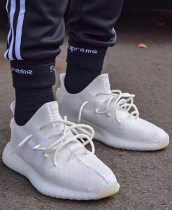 Close-up of white knit sneakers with laces on a person wearing black socks and track pants on a paved surface.