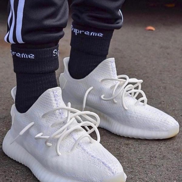 Close-up of white knit sneakers with laces on a person wearing black socks and track pants on a paved surface.