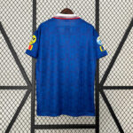 af3d186c | UAE SPORT STORE Back view of a blue football jersey hanging on a wooden hanger, featuring a red-and-white collar and subtle pattern on the fabric, with yellow patches on each sleeve against a lattice background.