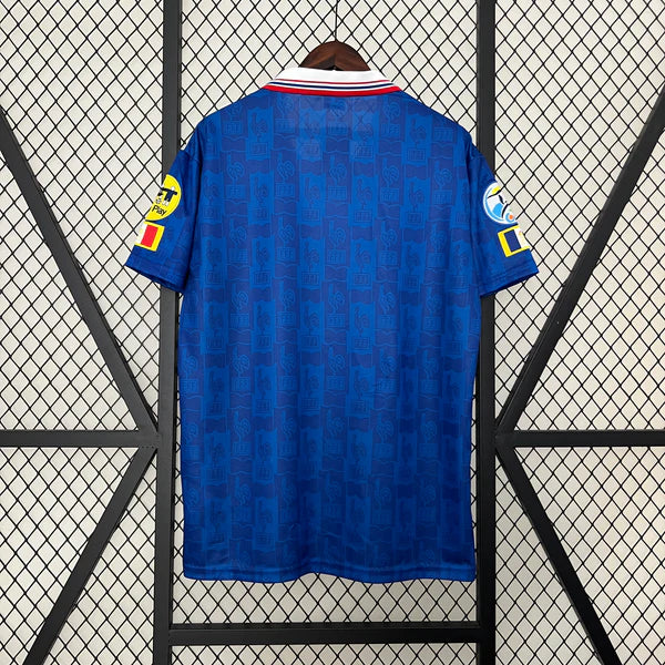 af3d186c | UAE SPORT STORE Back view of a blue football jersey hanging on a wooden hanger, featuring a red-and-white collar and subtle pattern on the fabric, with yellow patches on each sleeve against a lattice background.