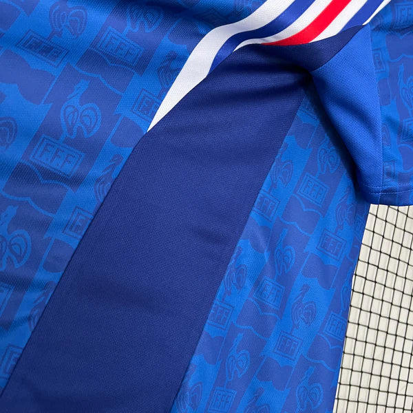 af7ced37 | UAE SPORT STORE Close-up of blue sports jersey fabric with a repeating emblem pattern and a red, white, and blue trim near a tennis net