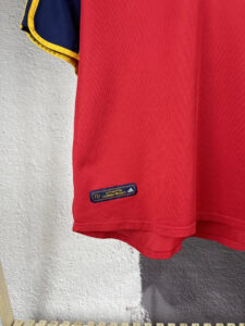 Red athletic shirt with a blue sleeve and yellow trim, featuring a small label reading 'AUTHENTIC LICENSED PRODUCT' and Adidas logo near the hem
