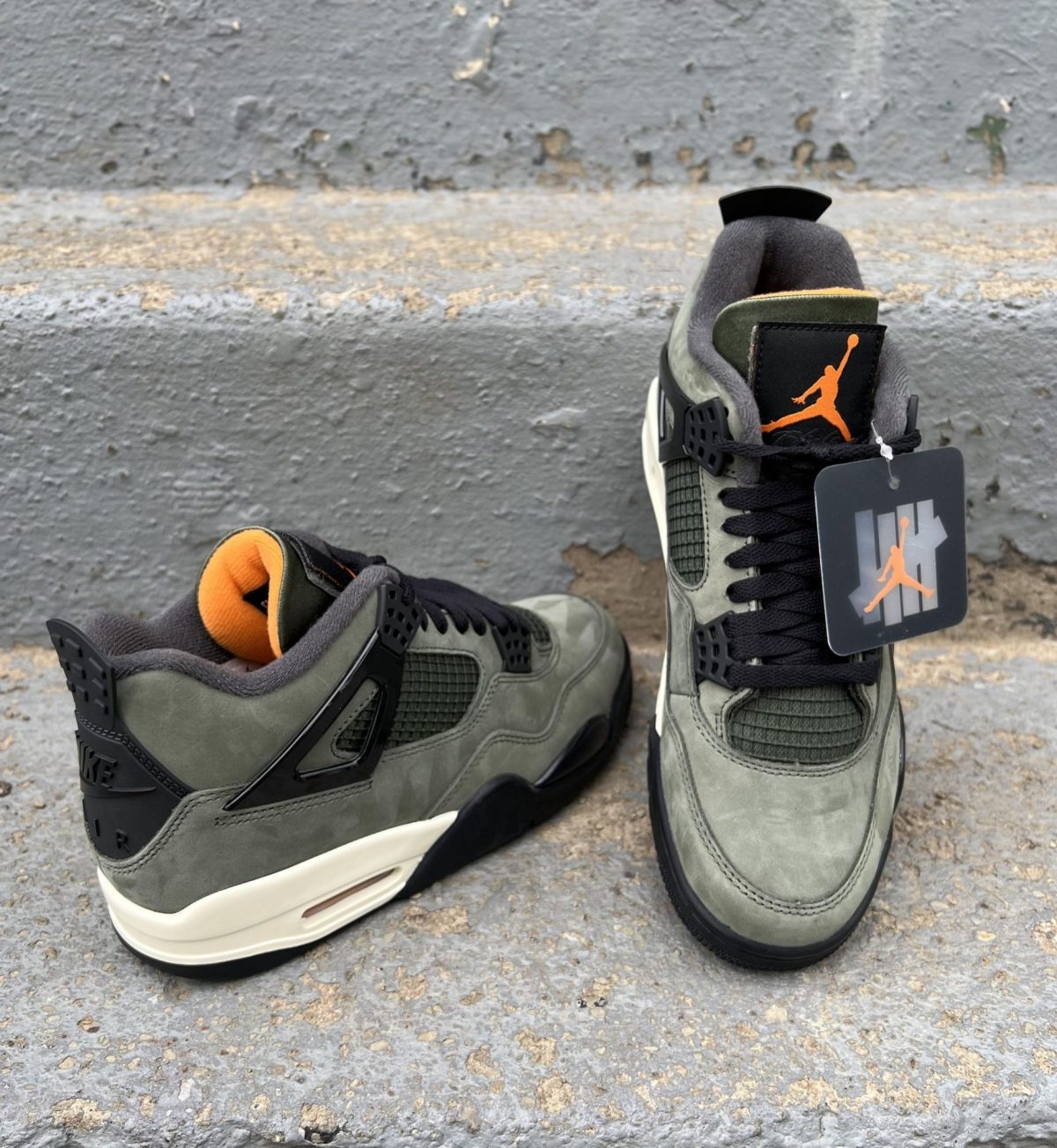 Top-down view of two olive-gray sneakers with orange lining, on a rough concrete surface; one shoe shows a Jordan tag hanging from the laces.