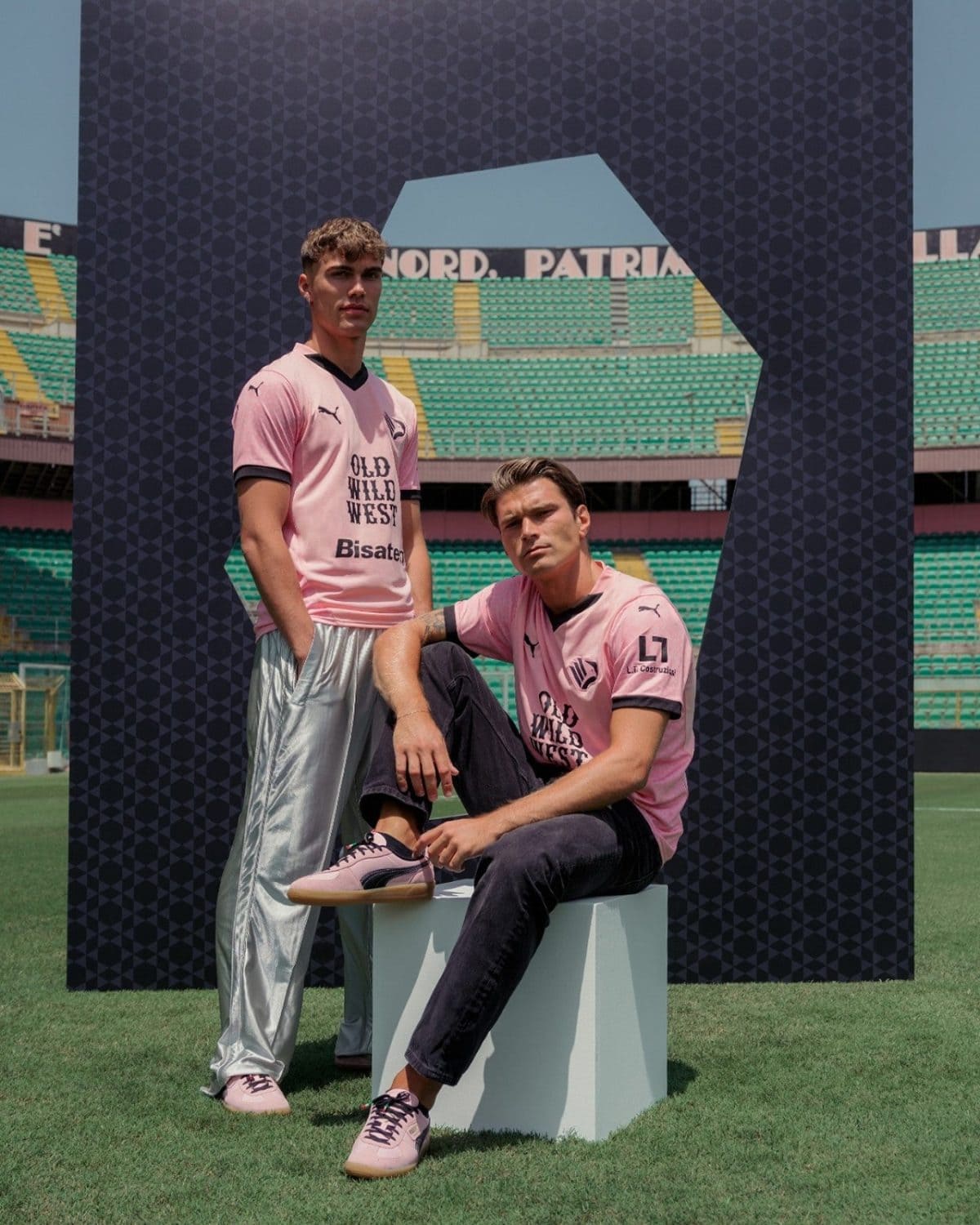 Two male models wear pink shirts and pants, posing in a stadium under a geometric arch on a grassy field.