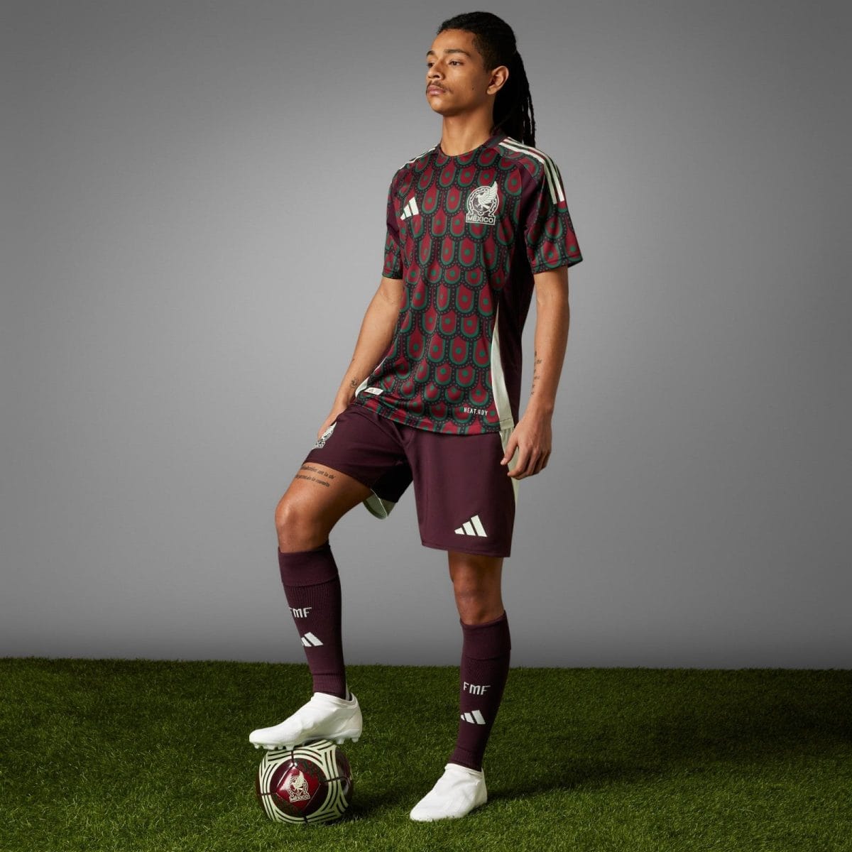 Male soccer player in maroon and teal Mexico jersey, standing with one foot on a patterned ball on grass field.