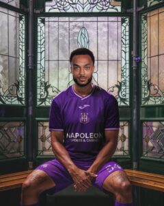 Portrait of a male athlete in a purple soccer kit seated in a stained-glass booth, facing the camera confidently.