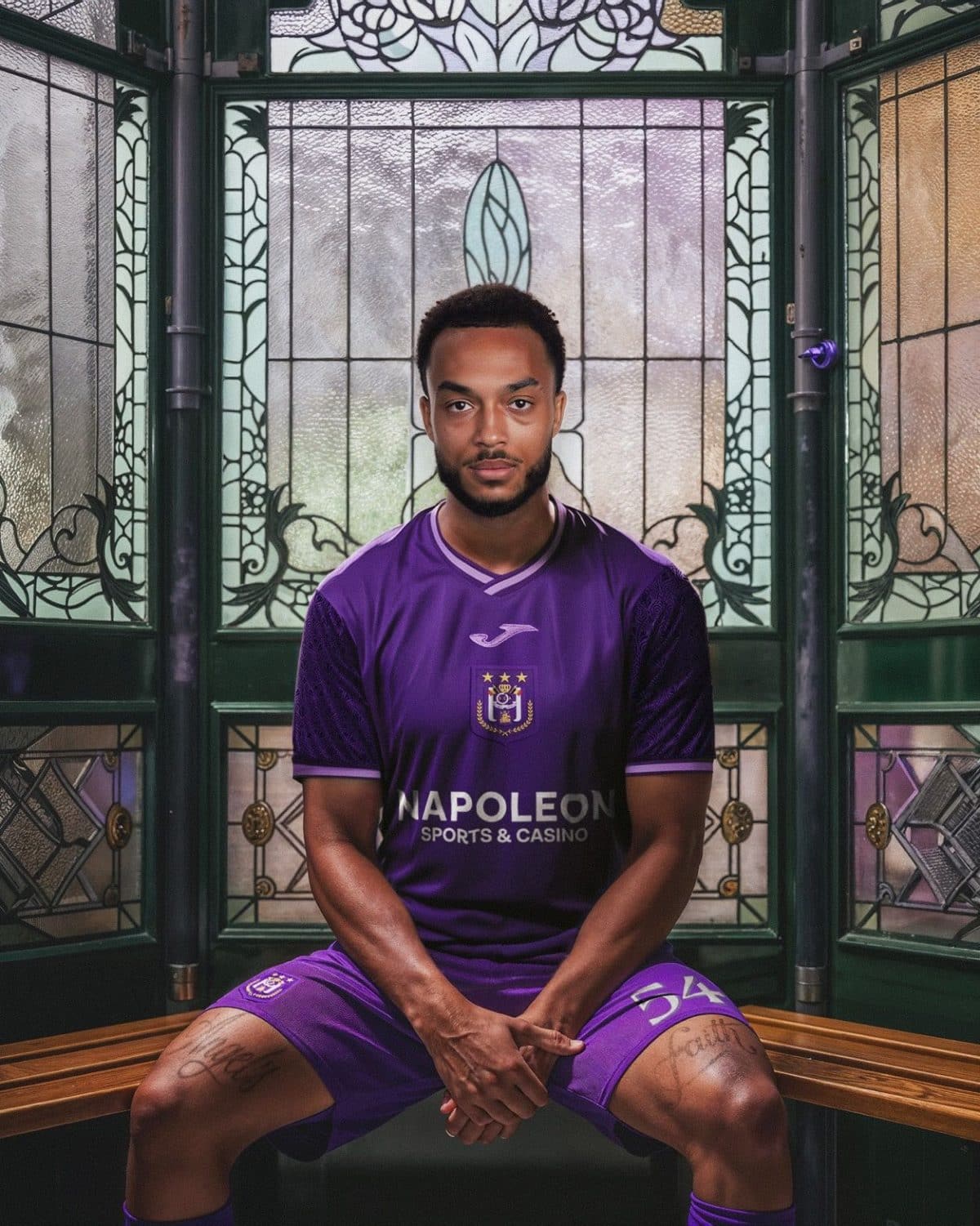 Portrait of a young man in a purple football kit sitting on a wooden bench inside a stained-glass booth, facing the camera. The jersey displays a crest and sponsor text.