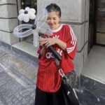 Woman in a red Adidas shirt stands outside a building, holding a large transparent balloon flower with a black stem and smiling.",
