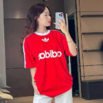 Woman taking a mirror selfie in a red Adidas-style jersey with white stripes and logo.