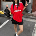 Person wearing a red Adidas jersey, black pleated mini skirt, white socks, and black shoes, posing with a black handbag outdoors.