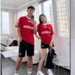 A couple stands in a bright room, both wearing matching red Adidas shirts and black shorts; the man takes a mirror selfie while the woman leans beside him with one hand on her hip.