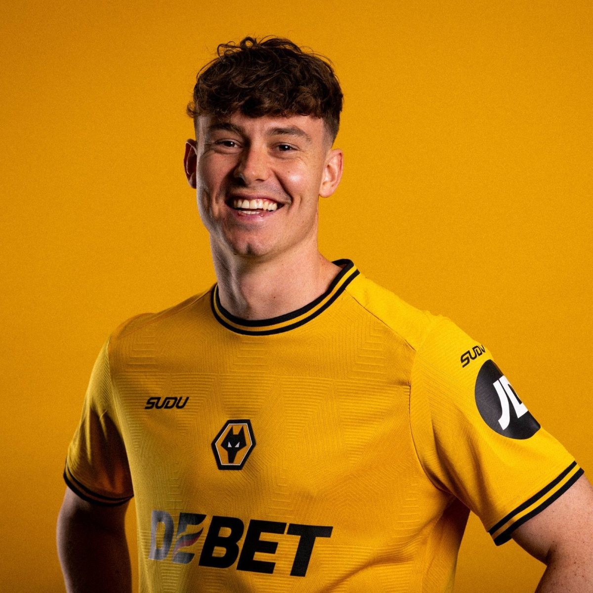 Smiling man in a yellow sports jersey with black trim, standing against a solid yellow background with a wolf emblem on the chest.