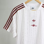 White Adidas jersey with vertical red pinstripes and maroon shoulder stripes on a wooden hanger, featuring a red Adidas trefoil logo and a small red cannon graphic on the chest.
