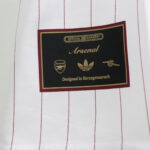 Gold-on-black Arsenal brand label with crest and cannon icons on white vertical-pinstriped fabric; reads 'Official Garnment' and 'Designed in Herzogenau r ch' (brand tag).