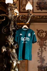 Teal AS Monaco soccer jersey hangs on an ornate chandelier statue in a dim, decorative room.