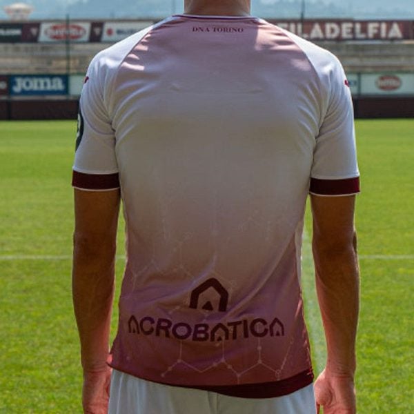 Athlete standing on a green sports field, back to camera, wearing a white-pink gradient jersey with the word 'ACROBATICA' on the lower back.