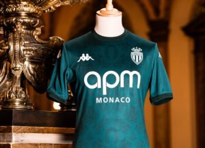 Teal AS Monaco football jersey on a mannequin, showing sponsor 'apm MONACO' and club crest against a ornate gold backdrop.