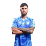 Athlete with short dark hair and beard standing with arms crossed, wearing a blue patterned sports jersey.