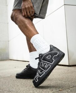 Person wearing black FILA sneakers with white detailing and large 'FILA' lettering, walking outdoors in white socks and gray shorts.