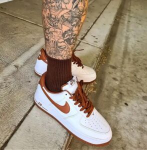 White Nike Air Force 1 sneakers with brown swoosh, laces, and sole, paired with dark brown socks and tattooed leg visible on a sidewalk edge.