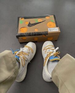 Top-down view of white sneakers with yellow laces and blue-yellow accents beside a Nike shoebox with orange circular designs on a dark floor.