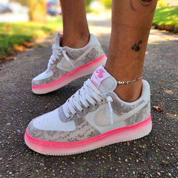 White sneakers with a bright pink sole on a person standing on a paved sidewalk; visible ankle tattoo and bracelet.
