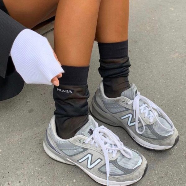 Gray New Balance sneakers with white laces on both feet, paired with black Prada socks, standing on a pavement.