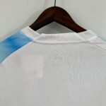 b1259ab9_79e0c5b4-c4f0-45a1-8d65-6e3ae7055f31 | UAE SPORT STORE Wooden clothes hanger holding a white shirt with light blue trim, against a plain wall background.