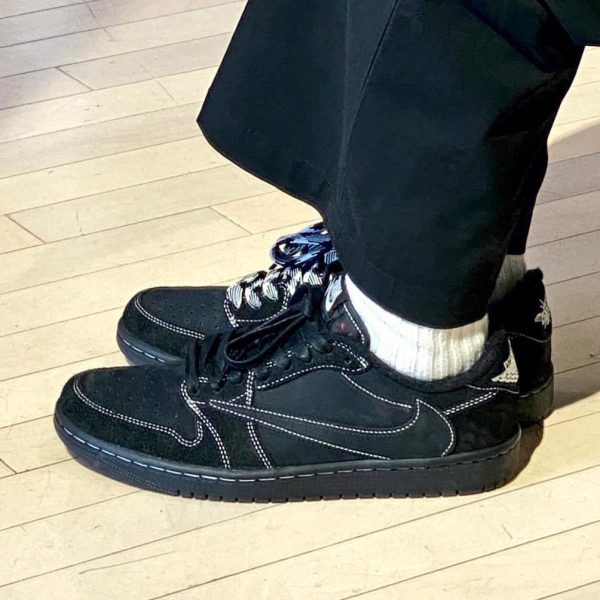 Black sneakers with white stitching and laces, worn with white socks on a light wooden floor.