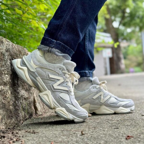 Person wearing light gray sneakers stepping onto a sidewalk next to a stone edge, jeans cuffs visible, greenery in the background