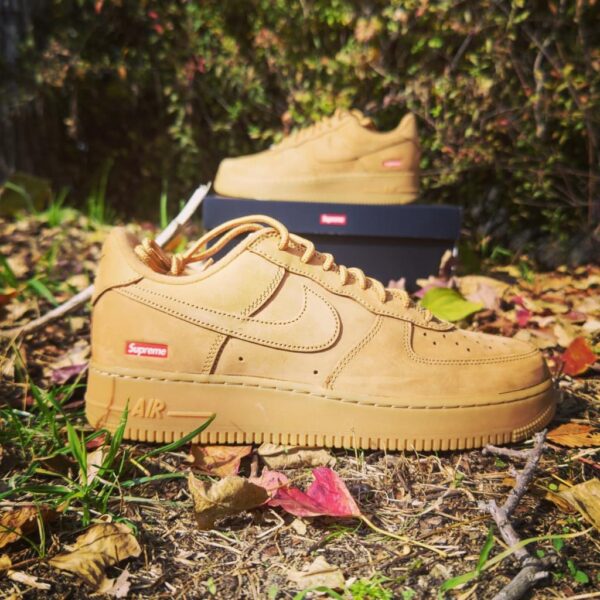 Tan Nike Air Force 1 sneaker with a red Supreme logo on the heel, outdoors among fallen leaves; shoebox in the background.