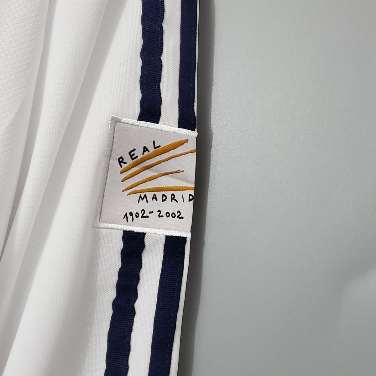 White fabric with a dark blue stripe and a small tag reading 'REAL MADRID 1902-2002', suggesting a commemorative scarf or ribbon.