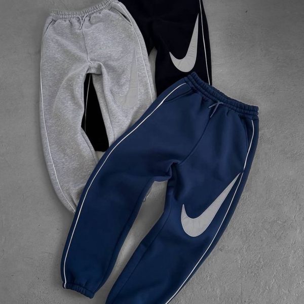 Three Nike jogger pants in grey, black, and navy with white piping and large swoosh logos laid on a grey surface.