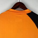 Orange shirt on a wooden hanger, showing the back neckline with a black sleeve panel against a light gray wall.