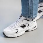 b2be3468-2e88-4260-bc47-78c468ca6ad7 | UAE SPORT STORE White New Balance running sneakers with black logo, laces tied, worn with blue jeans on a light gray floor