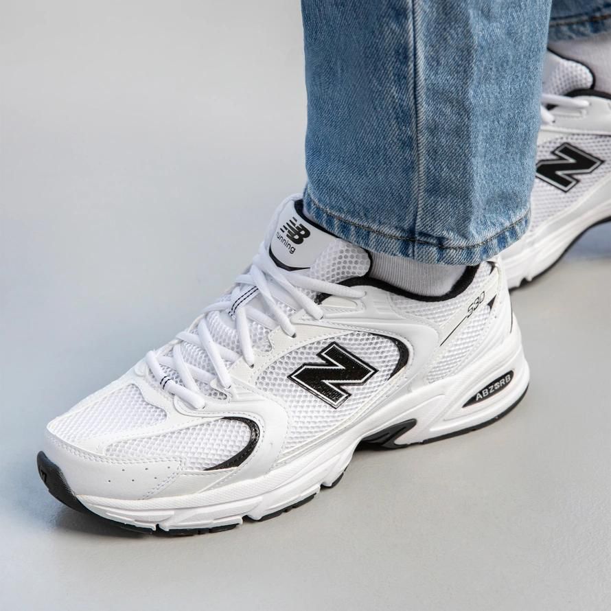 b2be3468-2e88-4260-bc47-78c468ca6ad7 | UAE SPORT STORE White New Balance running sneakers with black logo, laces tied, worn with blue jeans on a light gray floor