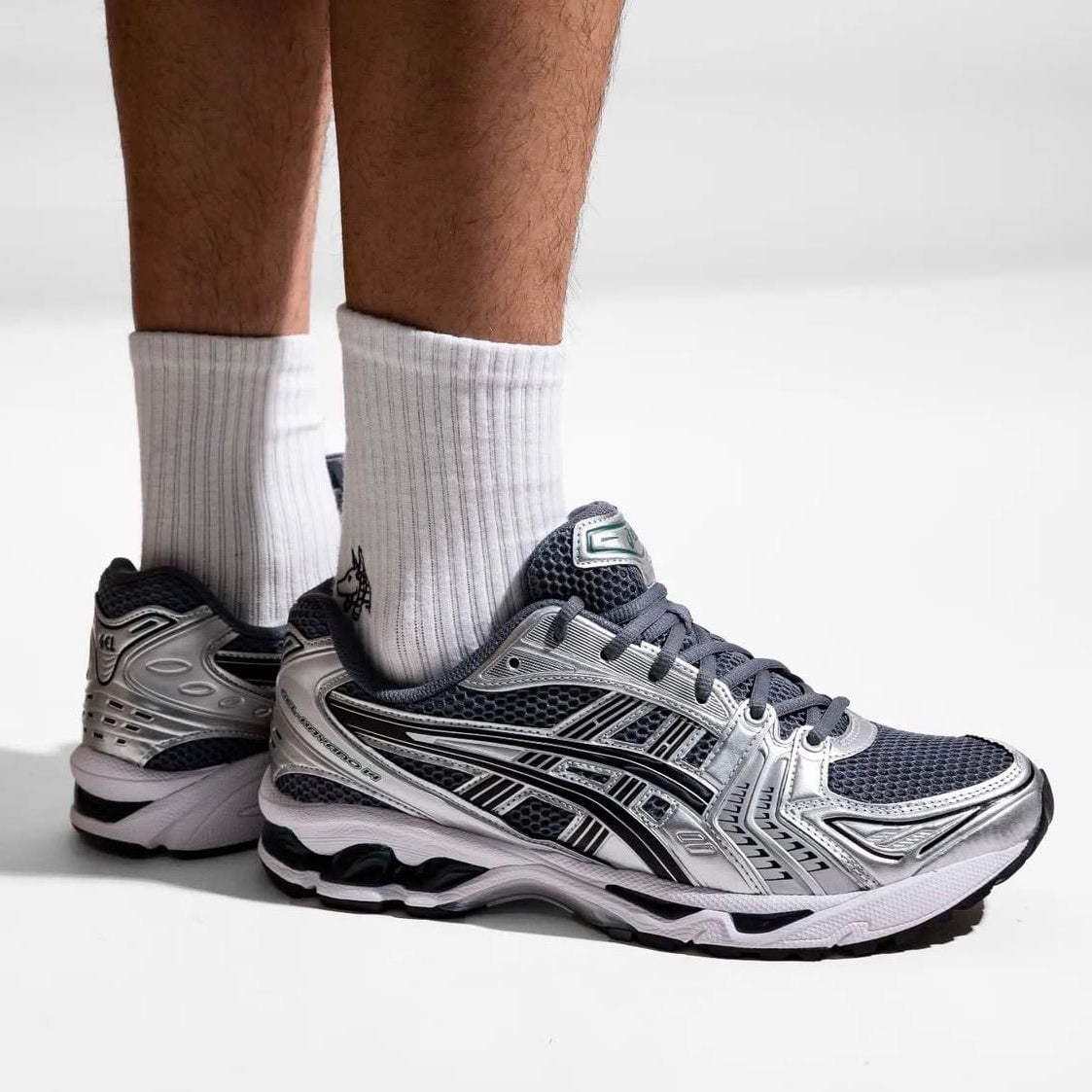 b3195f73-c064-41de-942c-e645bc5bf041 | UAE SPORT STORE Close-up of a person wearing white crew socks and silver/black running shoes standing on a white surface, showing the sneakers' details.