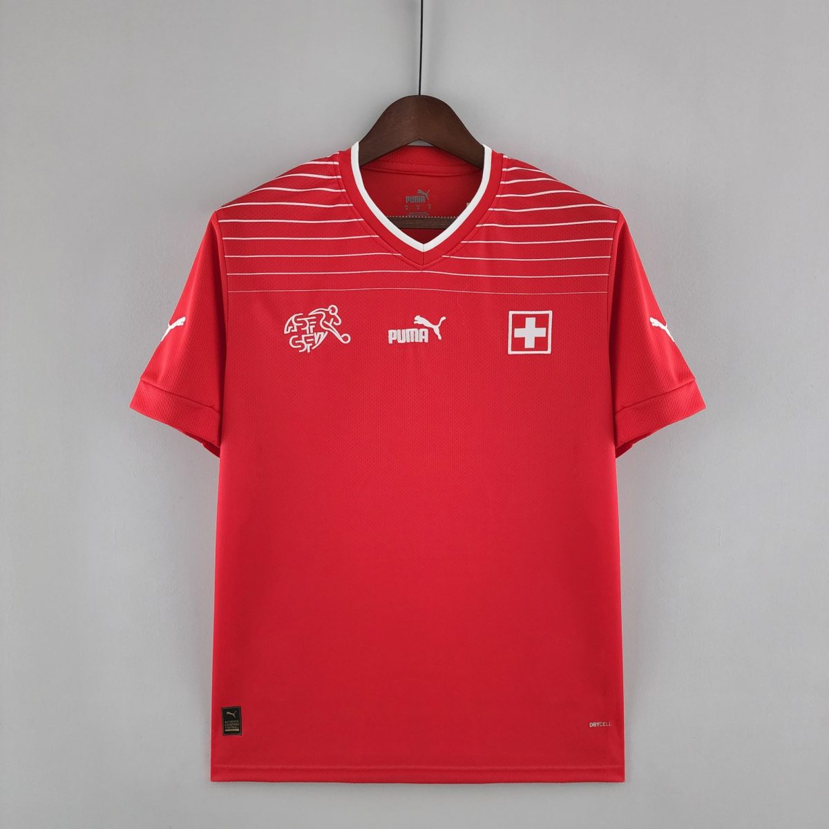 Red athletic jersey on a wooden hanger against a gray wall, with white horizontal stripes and a Swiss flag emblem on the chest.