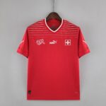 Red athletic jersey on a wooden hanger against a gray wall, with white horizontal stripes and a Swiss flag emblem on the chest.