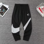 Black athletic joggers with a large white swoosh graphic on the left leg and a diagonal white stripe on the right leg, on a gray floor.