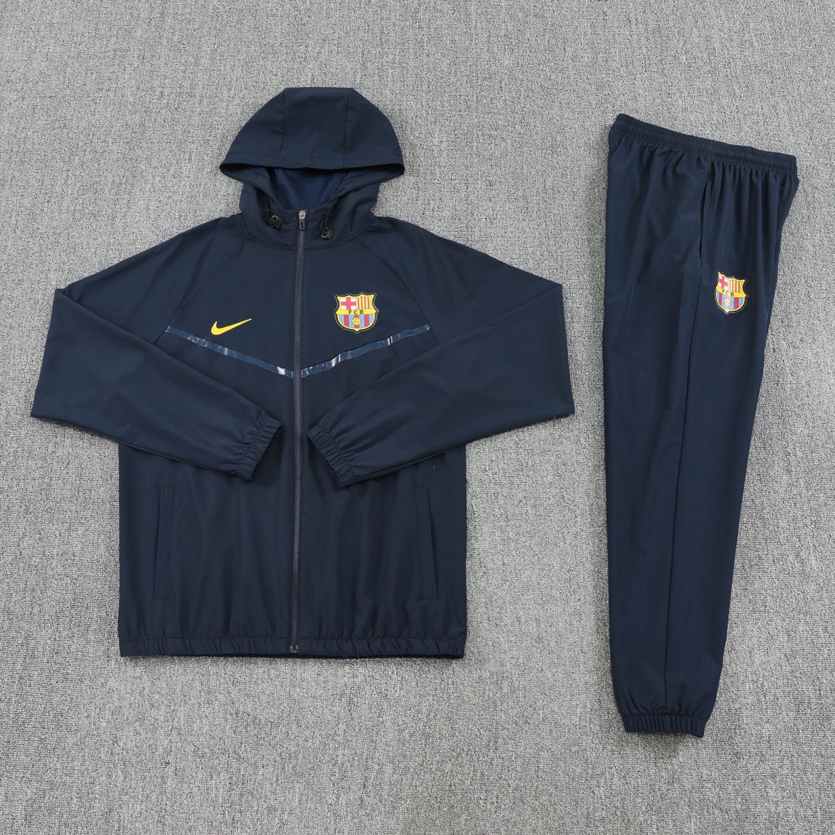 Navy blue FC Barcelona tracksuit jacket with crest and yellow Nike swoosh, laid flat next to matching pants on a gray carpet.
