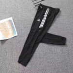 Black Adidas track pants with three white stripes, laid flat on a gray carpet beside a Supreme bag image accessory.