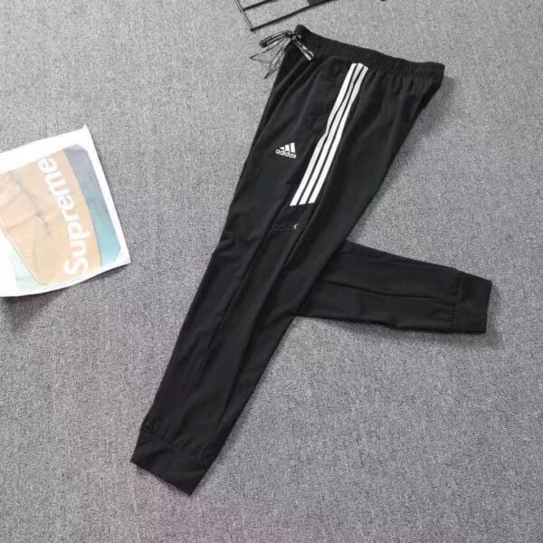 Black Adidas track pants with three white stripes, laid flat on a gray carpet beside a Supreme bag image accessory.