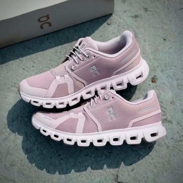 Light pink running shoes with chunky white soles resting on a concrete surface, a box in the background; brand logo visible on the side.