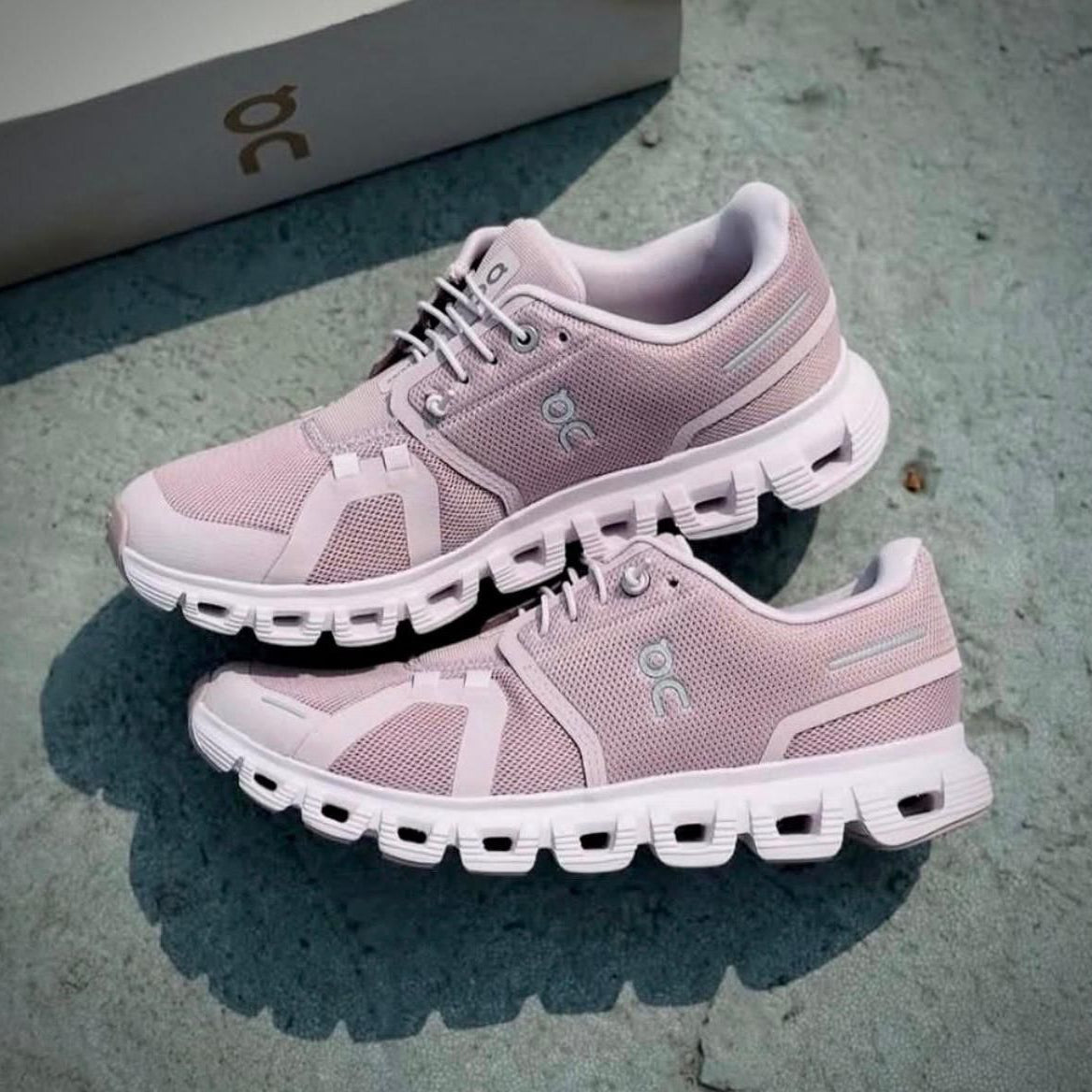 b576b1c1-90e7-46cd-aeb3-15a168d731cc Light pink running shoes with chunky white soles resting on a concrete surface, a box in the background; brand logo visible on the side.