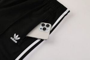 Black Adidas track pants with white side stripes and a zip pocket holding a smartphone.