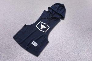 Navy sleeveless hoodie with hood and front pocket, white bull logo in a square on the chest, on a gray carpet.