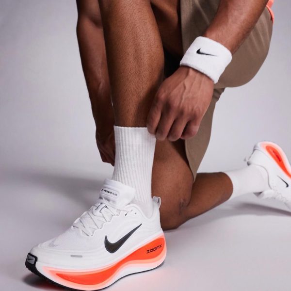Close-up of a runner wearing white Nike shoes with bright orange midsole, white socks, and beige shorts in a studio setting.