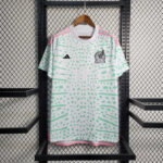 White Adidas Mexico soccer jersey with mint green abstract pattern and pink trim on a hanger against a metal grid background.